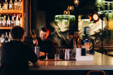 Bir barmen modern, şık bir barda sıcak ışıklandırma ve hoş bir ortam içeren bir kokteyl hazırlar. İçki şişeleri ve zarif cam eşyalar sofistike atmosferi güçlendirir..