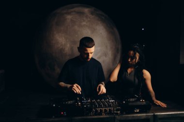 İki müzisyen, loş bir ortamda bir DJ istasyonunda sesi karıştırarak profesyonel ve sanatsal bir atmosferle canlı bir müzik sahnesi yaratıyorlar..