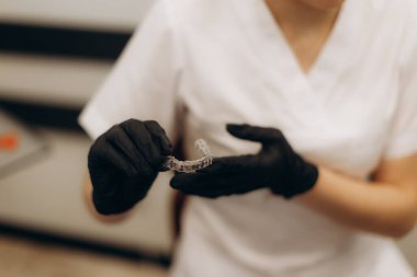 Siyah eldiven ve beyaz üniforma giyen bir dişçi görünmez bir diş düzenleyicisini gösteriyor. Yakın çekim sahnesinde profesyonel bir ortamda diş bakımı, hijyen ve modern ortodontik uygulamalar vurgulanıyor.