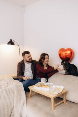 Bir çiftin kanepede birlikte suşinin tadını çıkardığı, kedileriyle bir anı paylaştığı sıcak bir ev ortamında aşkı, rahatlığı ve birlikteliği simgeleyen rahat bir ortam..