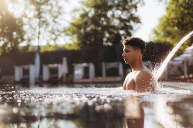 Genç adam lüks bir otel havuzunun ferahlatıcı suyunun keyfini çıkarıyor. Sırtından yumuşak bir şelale akıyor. Huzurlu ve rahatlatıcı bir atmosfer yaratıyor.