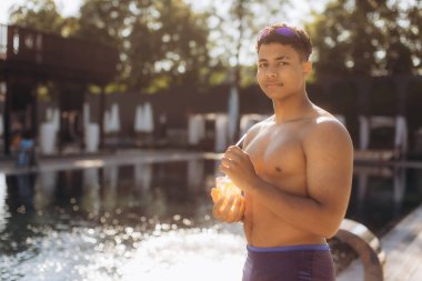 Lüks bir otelin havuz kenarında portakal suyu içen tişörtsüz genç adam yaz tatilinin tadını çıkarıyor.