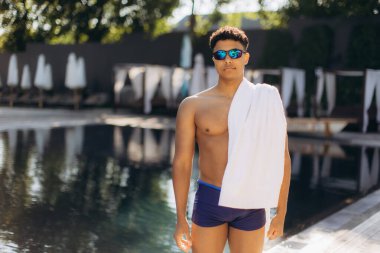 Genç erkek yüzücü, yaz güneşinin tadını çıkarırken, bir tatil köyü havuzunda antrenmandan sonra havluyla kurulanıyor.