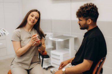Kadın dişçi, erkek hastaya takma diş modelini gösteriyor. Sandalyede oturuyor, diş sağlığı kliniğinde tedaviyi anlatıyor.