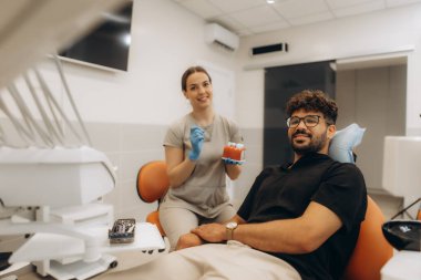 Dişlerinin anatomik modelini elinde tutan kadın dişçi, erkek hastaya klinikteki diş tedavisinin prosedürünü anlatıyor.