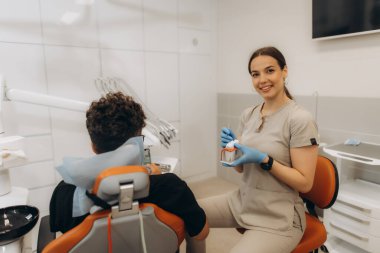 Kadın dişçi, anatomik bir model kullanırken diş prosedürünü açıklarken bir sandalyeye rahatça oturan bir hastayla meşgul oluyor.