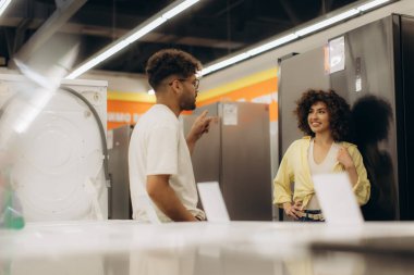 İki arkadaş, bir elektronik mağazasında modern cihazlar ve perakende ortamda tüketici etkileşimlerini vurgulayarak, bir yandan da bir elektronik mağazasında tartışırken, çamaşır makinelerini ve buzdolabını keşfediyorlar..