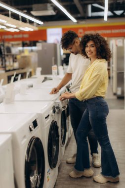 Gülümseyen bir çift çağdaş bir elektronik mağazasında çamaşır makinelerini keşfeder, modelleri değerlendirir, cihazlara odaklanmış bir alışveriş deneyimi sunar ve alışverişler sırasında karar verme mekanizmasını temsil eder..