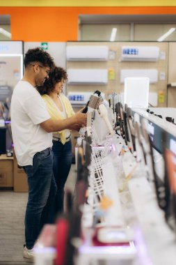 Bir çift, sergilenen çeşitli ürünlere göz atarak bir elektronik mağazasını keşfediyor. İhtiyaçlarına uygun en iyi seçenekleri arayarak eşyaları faiziyle incelerler..
