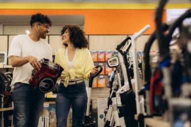 Modern bir elektronik mağazasında alışveriş yaparken farklı elektrik süpürgesi seçeneklerini araştıran mutlu çift..