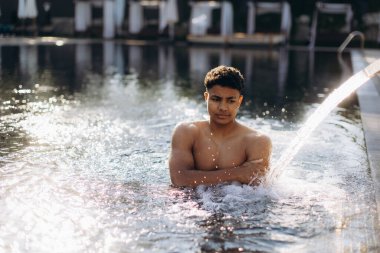 Genç adam lüks bir havuzda ferahlatıcı su jetinin tadını çıkarıyor, tatilinin sükunetini kucaklıyor.