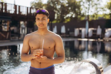 Elinde kokteyl bardağı tutan kaslı genç adam, lüks bir otel havuzunun yanında duruyor, rahatlama anının tadını çıkarıyor.
