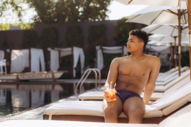 Genç adam serinletici bir kokteylin keyfini çıkarırken lüks bir tatil yerindeki havuzun kenarında güneşleniyor.