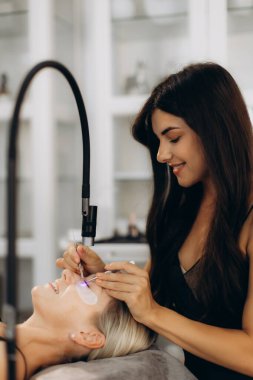 Güzellik uzmanı bir kadın müşteriye kirpik eklemek için kurşun ışığı kullanıyor, güzellik salonunda güzellik tedavisi uyguluyor.