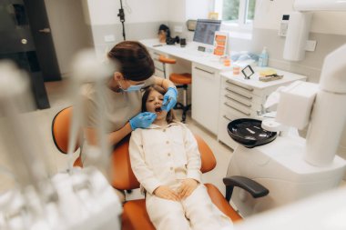 Maske takan ve eldiven takan kadın dişçi. Muayene sırasında dişçi koltuğunda oturan genç bir kızın dişlerini dikkatlice inceliyor.