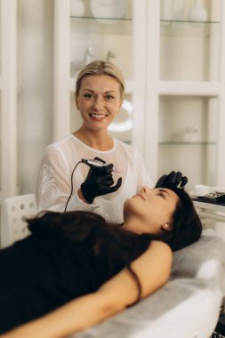 Müşteri kanepede güzellik yaratırken, kalıcı makyaj prosedürü için kaşına odaklanan bir güzellik uzmanı.