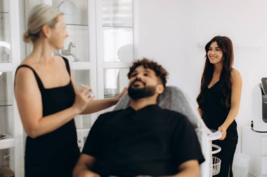 Genç bir adam kadın bir kozmetik uzmanından estetik ameliyatı olurken diğer bir bayan asistan beyaz bir güzellik kliniğinin yakınında duruyor.