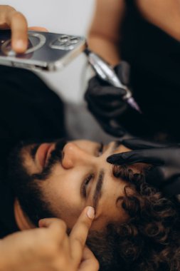 Profesyonel güzellik uzmanı bir adamın kaşlarına kalıcı makyaj uyguluyor, güzelliği arttırıyor ve tımar ediyor.