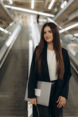 Kendine güvenen genç profesyonel dizüstü bilgisayarını tutarak yürüyen merdivenin yanında duruyor. Minimalizm ve çağdaş tasarım ile dengeli ve modern bir iş ortamı çiziyor..