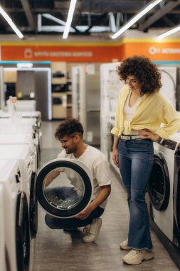 Bir çift, modern bir elektronik mağazasında tüketici tercihleri ve teknolojinin sergilendiği yeni bir çamaşır makinesi seçeneklerini araştırıyor. Parlak çevre, elverişli ve yenilikçi ev aletlerini vurguluyor.