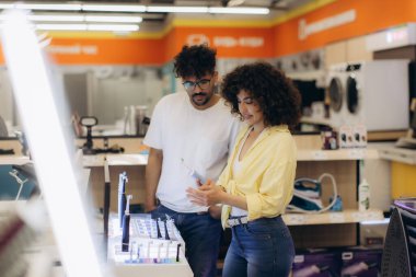 Bir erkek ve bir kadın, aydınlık ve modern bir mağazada elektronik aletleri inceleyerek alışveriş deneyiminin tadını çıkarıyorlar. Bir çift diş fırçasını seçer.