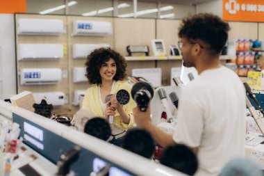 İki kişi, modern bir elektronik mağazasında saç kurutma makinelerine göz atarak, en iyi ürün seçeneğine karar vermek için arkadaşça bir anı paylaşıyorlar..