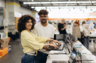 Neşeli bir çift, bir elektronik mağazasında dizüstü bilgisayarlar için birlikte alışveriş yapar, seçenekleri araştırır ve rahatlatıcı, çağdaş bir perakende ortamda alışveriş deneyiminin tadını çıkarır..