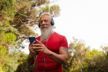 Beyaz sakallı son sınıf öğrencisi dışarıda koşuyor, telefonu tutuyor ve müziği seçiyor.