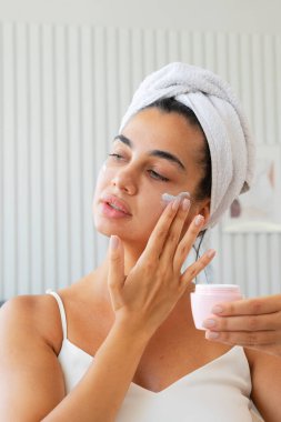Oturma odasında oturmuş banyo yaptıktan sonra nemlendirici yüz kremi sürerken kafasına havlu sarılmış neşeli, sevimli genç bir kız. Cilt bakımı ve güzellik kavramı