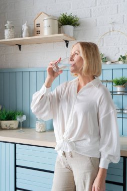 Gülümseyen bir kadın, şık bir mutfakta taze sebzelerle çevrili ferahlatıcı bir bardak su tutar.