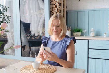 Mutfakta kahve içen ve cep telefonundaki mesajları kontrol eden yaşlı bir kadın.