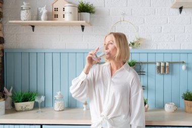 Gülümseyen bir kadın, şık bir mutfakta taze sebzelerle çevrili ferahlatıcı bir bardak su tutar.