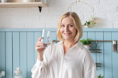 Gülümseyen bir kadın, şık bir mutfakta taze sebzelerle çevrili ferahlatıcı bir bardak su tutar.