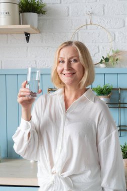 Gülümseyen bir kadın, şık bir mutfakta taze sebzelerle çevrili ferahlatıcı bir bardak su tutar.