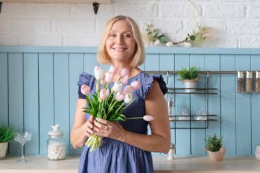 Güzel bir buket pembe ve beyaz laleyi sıcacık mutfağında tutarken neşeli bir kadın mutlulukla ışıldıyor.