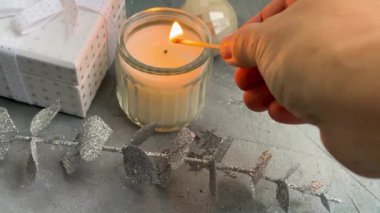 Sakin bir tatil düzeninde bir mum, zarif bir sabun ve desenli bir arka planda şık gümüş yapraklar bulunur.