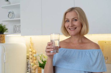 Gülümseyen bir kadın, şık bir mutfakta taze sebzelerle çevrili ferahlatıcı bir bardak su tutar.