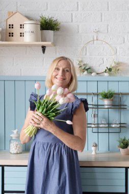 Güzel bir buket pembe ve beyaz laleyi sıcacık mutfağında tutarken neşeli bir kadın mutlulukla ışıldıyor.