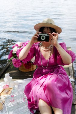 Pembe bir elbise ve uyumlu bir şapka giyen şık bir kadın, sakin bir su cesedinin yanında fotoğraf çekmekten zevk alır.