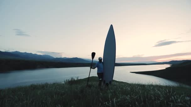 哈萨克斯坦落日时分 一个留着胡子的男人站在湖畔 手里拿着桨和苏打水冲浪板 户外休闲活动 自然是慢动作 — 图库视频影像