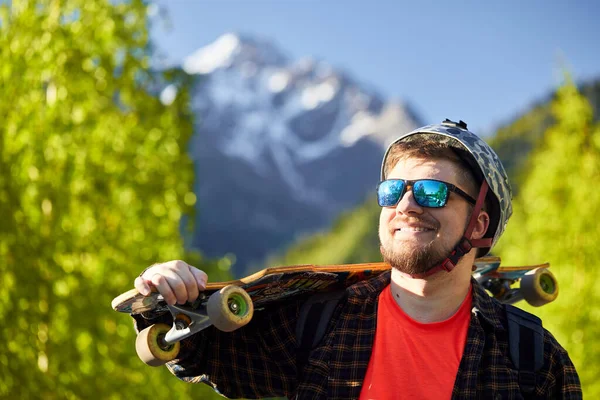 带着太阳镜 头戴安全帽 快乐胡子男子滑板的画像 背景是山中的长板 — 图库照片