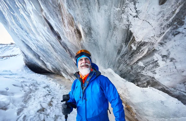 Portrait of smiling old bearded man with action camera in blue costume near ice cave wall at glacier in mountain valley covered with snow in Almaty, Kazakhstan