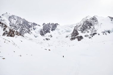 Sırt çantalı küçük adam güzel dağlarda kar tepesinde yürüyor Almaty, Kazakistan 'daki bulutlu gökyüzüne karşı. 