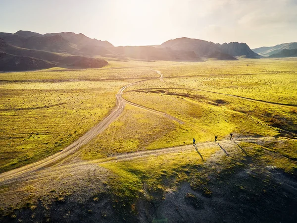 空中无人驾驶飞机俯瞰着三个运动员在尘土飞扬的乡间道路上飞奔 他们在青草丛中 夕阳西下 群山丛生 哈萨克斯坦南部草原上的小径 — 图库照片