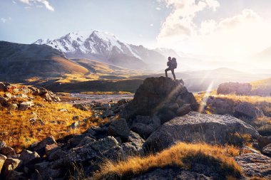 Büyük sırt çantalı küçük adam, Orta Asya, Kazakistan 'da günbatımı bulutlu arka planda karlı zirveleri olan dağ vadisine tırmanıyor.