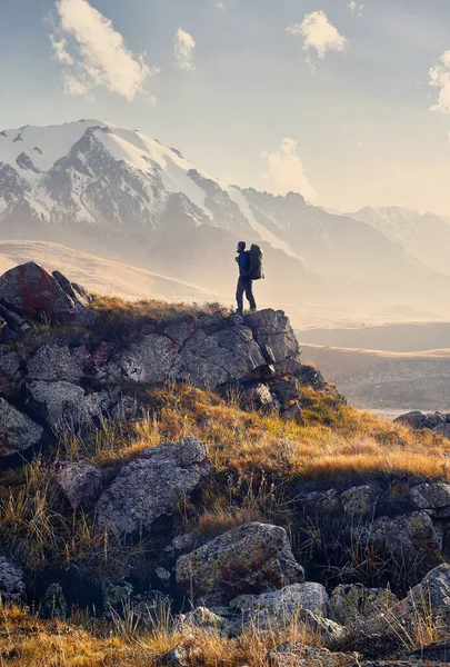 大きなバックパックを持つ小さな男の観光客は 中央アジア カザフスタンの日没の曇りの空の背景で雪のピークと山の谷に登る歩いている — ストック写真