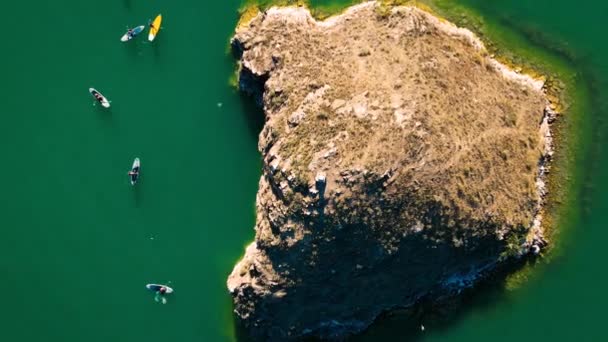 空中スタンドアップパドルボードSupで漕ぐ人々のグループのトップダウンドローンショット カザフスタンのカチャガイ湖に海鳥のいる小さな岩の島日没時 — ストック動画