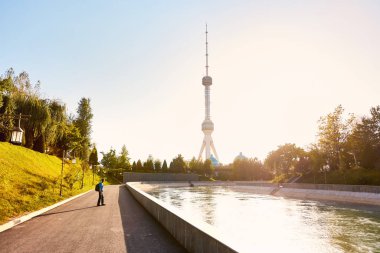Sırt çantalı turist, Özbekistan 'da gün batımında parktaki nehrin yanındaki Taşkent Televizyon Kulesi' ne bakıyor.