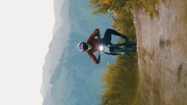 Dağ bisikletçisinin dikey videosu, kasklı ve elektrikli motosikletle dağ yolunda tek tekerleğinde parlak bir ışık..