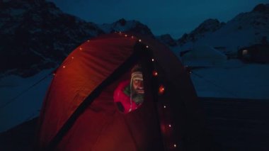 Kırmızı ceketli bir adam kamp çadırında oturuyor. Parlayan Noel çelengi ışıklarıyla pencereden bakıyor. Gece vakti kış dağları.